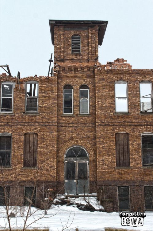 Abandoned brick building with a tower, broken windows, and a snow-covered ground, showcasing decay and neglect.