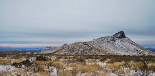 A snowy mountain landscape with a prominent peak and a cloudy sky, surrounded by sparse vegetation.