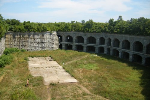 Abandoned stone structure surrounded by overgrown grass and trees, with arched openings and a clear blue sky.
