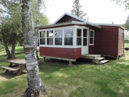 A small red cabin with a porch, surrounded by trees and a grassy area, featuring a picnic table nearby.