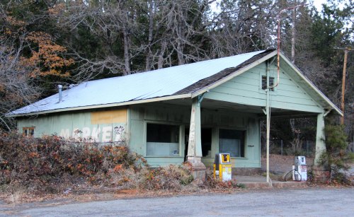 Abandoned green building with a metal roof, overgrown vegetation, and old gas pumps in front.