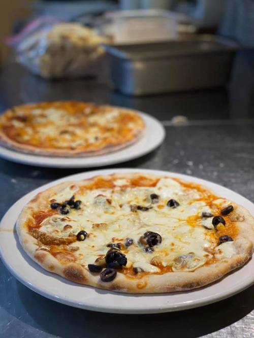 Two pizzas on white plates, topped with cheese and black olives, on a stainless steel countertop.