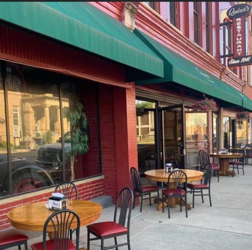 Exterior of a restaurant with green awnings, outdoor tables, and a brick facade on a sunny day.