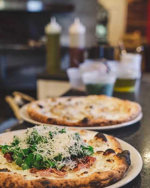 Two pizzas on plates, one topped with greens and cheese, the other in the background, with condiments visible.