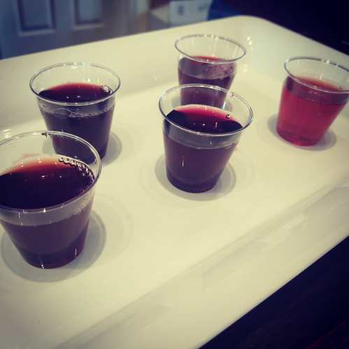 Five small cups with different colored liquids on a white tray, set against a blurred background.