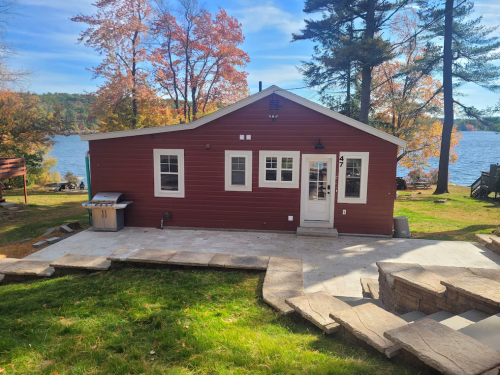 A red cabin by a lake, surrounded by trees with autumn foliage, featuring a stone patio and grill.