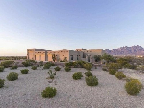 A modern adobe-style house surrounded by desert landscape and mountains in the background during sunset.