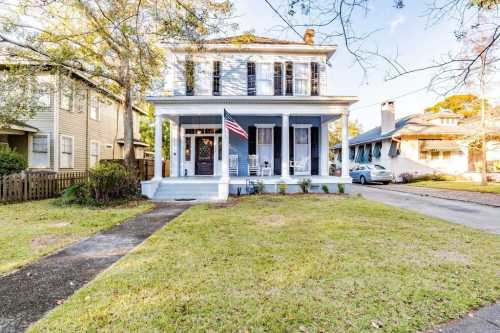 A charming two-story house with a front porch, American flag, and well-maintained lawn in a residential neighborhood.