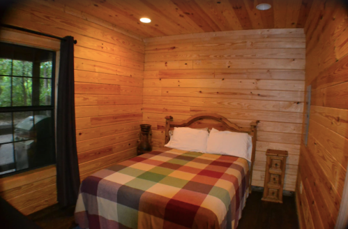Cozy bedroom with wooden walls, a double bed covered in a colorful plaid blanket, and a bedside table.