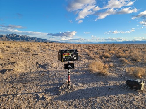 A weathered mailbox with stickers stands in a barren landscape under a blue sky with scattered clouds and distant mountains.