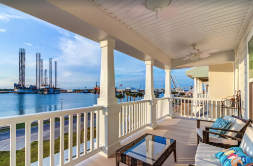 A scenic view from a balcony overlooking a harbor with drilling rigs and a calm waterway under a blue sky.