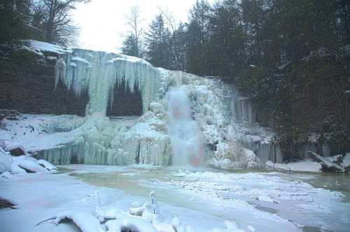 A frozen waterfall surrounded by snow-covered rocks and trees, creating a serene winter landscape.