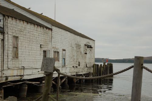 Weathered building on stilts by the water, with a cloudy sky and distant figures standing on a pier.