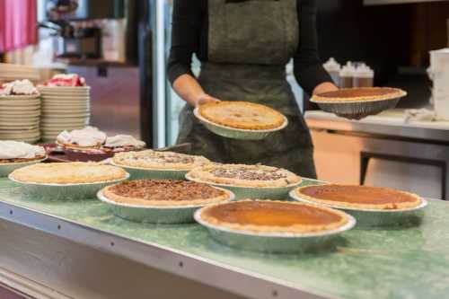 A person holds two pies over a table with various pies displayed, set in a kitchen or dining area.