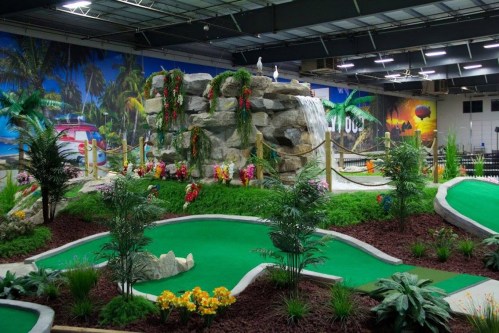 Indoor mini-golf course featuring a waterfall, lush greenery, and colorful flowers against a tropical mural backdrop.