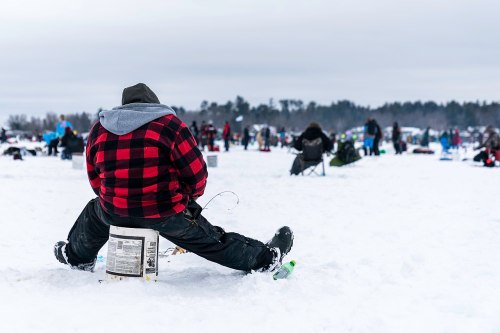 A person in a red and black checkered jacket sits on a bucket in the snow, surrounded by others ice fishing.