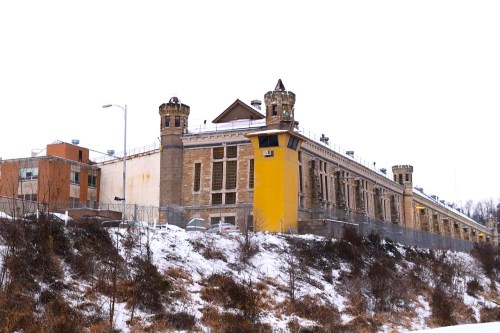 Historic prison building with towers, surrounded by snow and a fence, set against a gray winter sky.