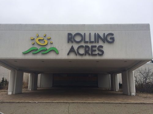Sign for "Rolling Acres" with a sun and wave design, set against a gray building under overcast skies.