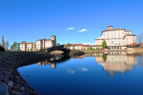 A serene lakeside view featuring buildings and a bridge under a clear blue sky. Reflections shimmer on the water.