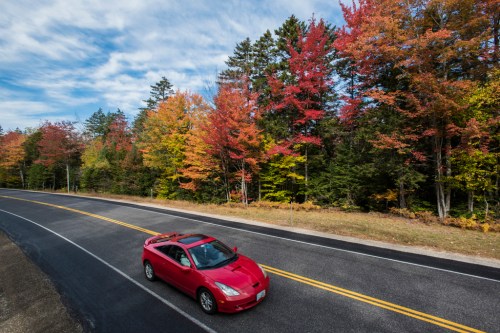 A red car drives along a winding road lined with vibrant autumn trees in shades of red, orange, and yellow.