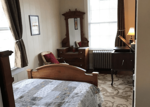 Cozy bedroom featuring a bed, dresser, mirror, and a warm lamp by the window, with patterned carpet and curtains.
