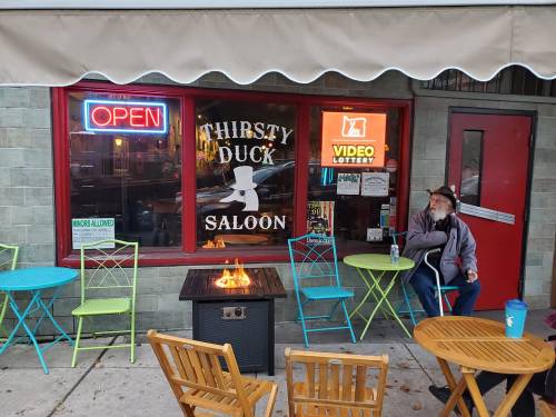 A cozy saloon with colorful outdoor seating, a fire pit, and a sign reading "Thirsty Duck Saloon" in the window.