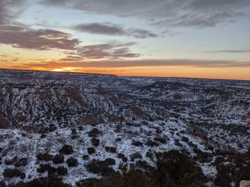 A scenic view of a snowy canyon at sunset, with layered hills and a colorful sky in the background.