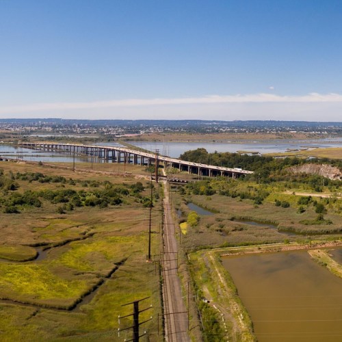 Aerial view of a river with a bridge, surrounded by green fields and trees under a clear blue sky.