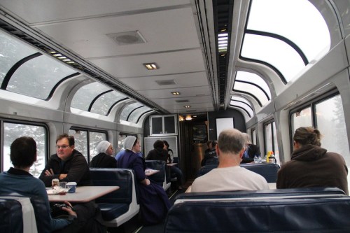 Interior of a train dining car with passengers seated at tables, large windows showing a snowy landscape outside.