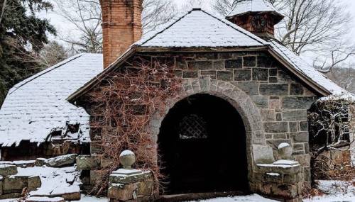 A stone structure with an arched entrance, covered in snow, surrounded by trees and a chimney in a winter landscape.
