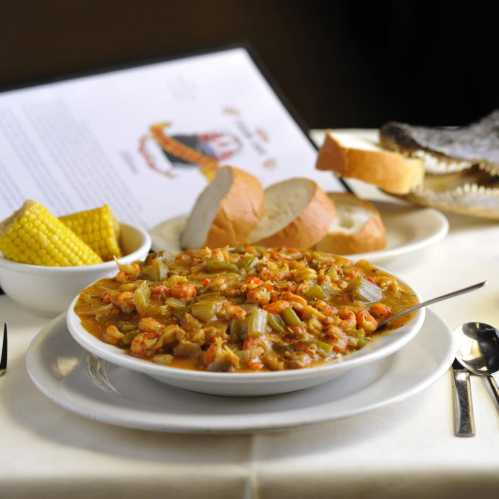 A bowl of crawfish stew with celery, served with bread, corn on the side, and a menu in the background.