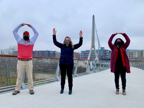 Three people on a bridge, wearing masks, standing with arms raised in a "O" shape against a cloudy sky.