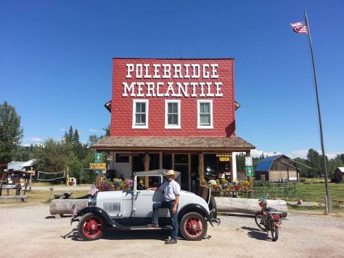 A vintage car parked in front of Polebridge Mercantile, with a man standing beside it and an American flag flying.