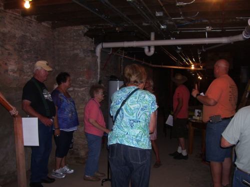 A group of people stands in a dimly lit basement, listening to a speaker near exposed pipes and stone walls.