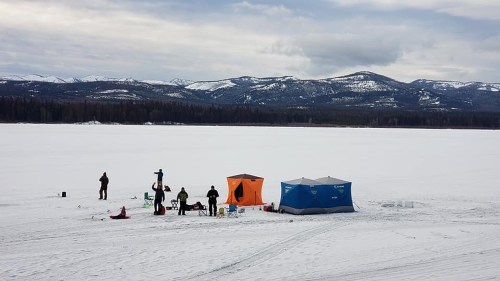 A snowy landscape with people ice fishing near colorful tents, surrounded by mountains under a cloudy sky.