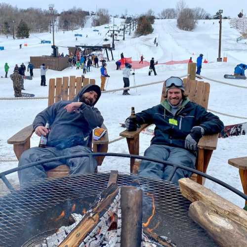 Two men relax in chairs by a fire pit, enjoying drinks, with a snowy ski resort and people in the background.