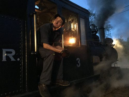 A train engineer sits in the cab of a steam locomotive, illuminated by a warm light, with smoke billowing around.