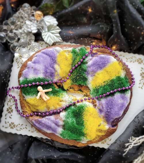 A colorful King Cake decorated with purple, green, and yellow sugar, adorned with a small figurine and beads.