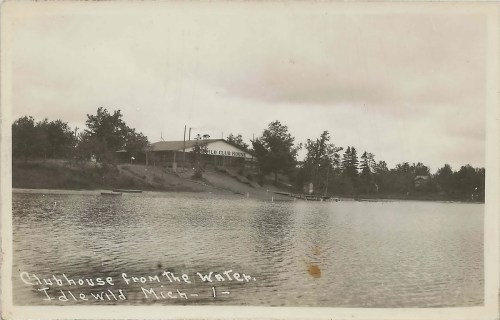 Black and white photo of a lakeside clubhouse surrounded by trees, with a calm water surface in the foreground.