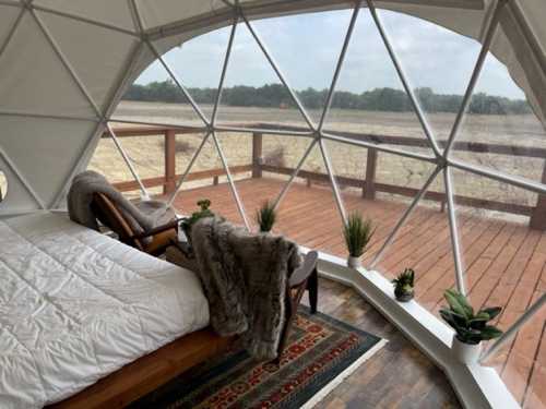 Cozy interior of a geodesic dome with a bed, chair, and plants, overlooking a wooden deck and open landscape.