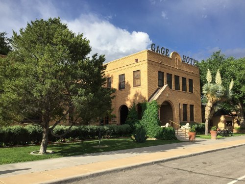 Historic Gage Hotel with brick facade, greenery, and palm trees under a partly cloudy sky.
