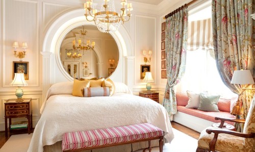 Elegant bedroom featuring a large bed, decorative curtains, a round mirror, and a cozy seating area by the window.