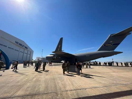 A military aircraft parked near a hangar, with a crowd of people gathered around on a sunny day.