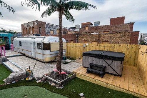 A vintage trailer beside a hot tub, surrounded by a wooden deck, palm tree, and colorful landscaping.