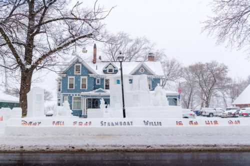 A snow sculpture in front of a blue house, featuring the words "Snow Man from Steamboat Willie is an old man."