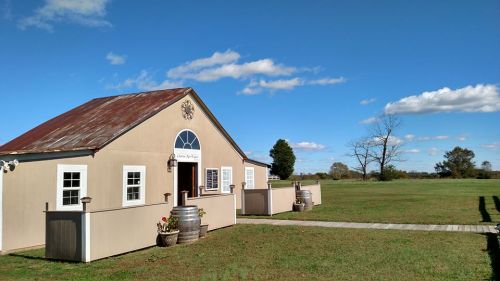 A charming building with a red roof, surrounded by green grass and a clear blue sky, featuring a pathway and decorative barrels.