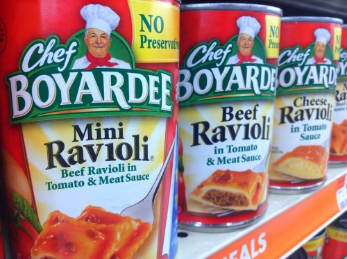 Cans of Chef Boyardee Mini Ravioli, Beef Ravioli, and Cheese Ravioli in tomato and meat sauce on a store shelf.