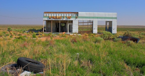 Abandoned building in a grassy landscape, with broken windows and a tire in the foreground under a clear blue sky.