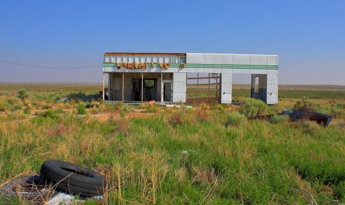 Abandoned building in a grassy landscape, with tires and overgrown vegetation under a clear blue sky.