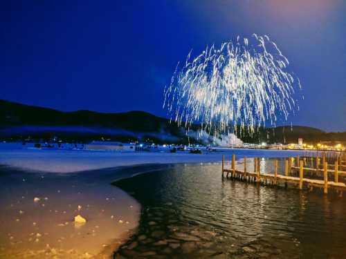 Fireworks illuminate the night sky over a snowy landscape and calm water, with a dock in the foreground.